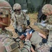 U.S. Army soldiers are conducting a field medical training exercise, treating a simulated casualty on a litter in an outdoor environment near a military tent. Multiple personnel are performing coordinated medical tasks including airway management and documentation. Soldiers are wearing OCP (Operational Camouflage Pattern) uniforms with unit patches visible, including a 1st Cavalry Division patch.