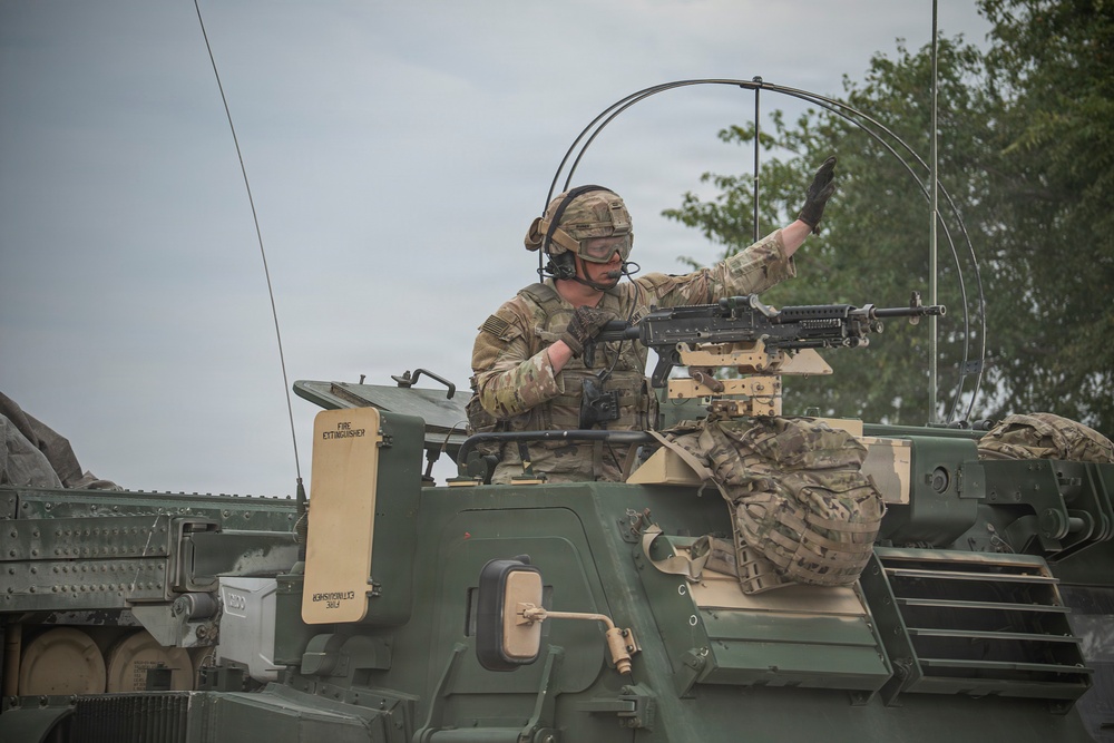A U.S. Army soldier in full combat gear is positioned in the hatch of an armored military vehicle, raising one hand in a signaling gesture. The soldier is equipped with a helmet, communications headset, protective eyewear, and a mounted machine gun is visible on the vehicle. The image appears to depict a training exercise or operational movement.
