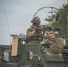 A U.S. Army soldier in full combat gear is positioned in the hatch of an armored military vehicle, raising one hand in a signaling gesture. The soldier is equipped with a helmet, communications headset, protective eyewear, and a mounted machine gun is visible on the vehicle. The image appears to depict a training exercise or operational movement.
