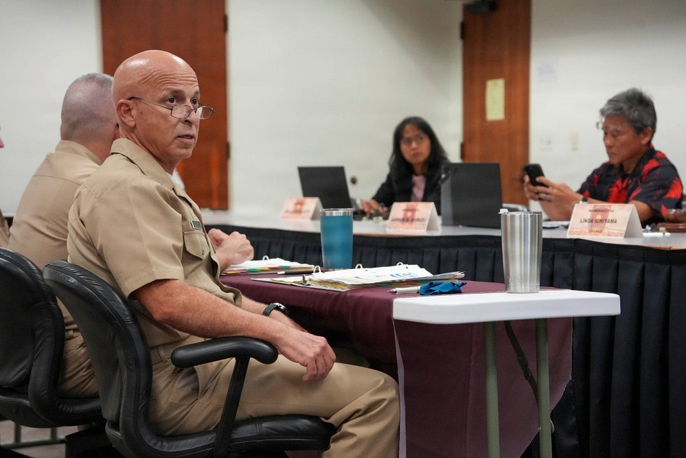 A meeting or briefing is taking place in a conference room setting. A bald man wearing military khaki uniform with glasses is seated at a table and appears to be speaking or listening. Other attendees, including civilians, are visible in the background seated around a larger table with name placards.