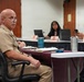 A meeting or briefing is taking place in a conference room setting. A bald man wearing military khaki uniform with glasses is seated at a table and appears to be speaking or listening. Other attendees, including civilians, are visible in the background seated around a larger table with name placards.