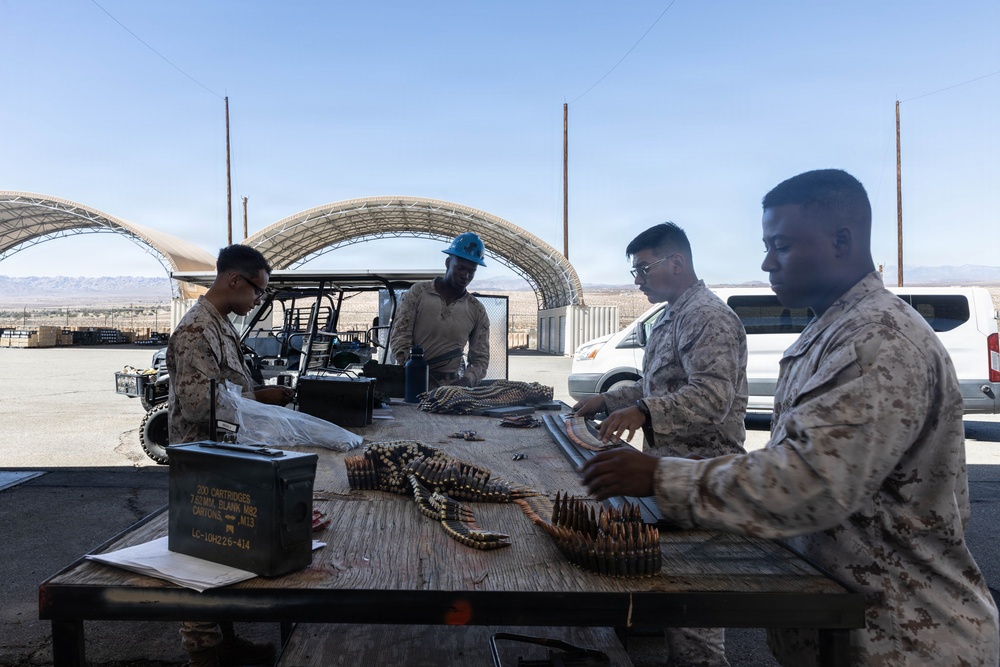Four military personnel in MARPAT (Marine Corps) camouflage uniforms are working at an outdoor table handling ammunition and equipment. They appear to be sorting, loading, or inspecting linked ammunition belts and magazines at what appears to be a desert military installation. An ammunition can labeled '200 Cartridges 7.62MM Blank M82' is visible on the table.