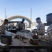 Four military personnel in MARPAT (Marine Corps) camouflage uniforms are working at an outdoor table handling ammunition and equipment. They appear to be sorting, loading, or inspecting linked ammunition belts and magazines at what appears to be a desert military installation. An ammunition can labeled '200 Cartridges 7.62MM Blank M82' is visible on the table.