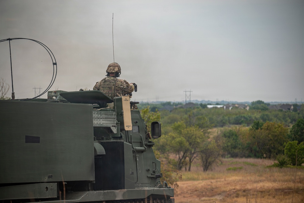 A soldier in camouflage uniform and helmet is positioned in the open hatch of a large military armored vehicle, facing away from the camera toward an open landscape. The vehicle appears to be a tracked armored personnel carrier or self-propelled artillery system painted in olive drab green. The setting appears to be a semi-arid training or operational area with scattered trees, dry grass, and power lines visible in the background.