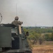A soldier in camouflage uniform and helmet is positioned in the open hatch of a large military armored vehicle, facing away from the camera toward an open landscape. The vehicle appears to be a tracked armored personnel carrier or self-propelled artillery system painted in olive drab green. The setting appears to be a semi-arid training or operational area with scattered trees, dry grass, and power lines visible in the background.
