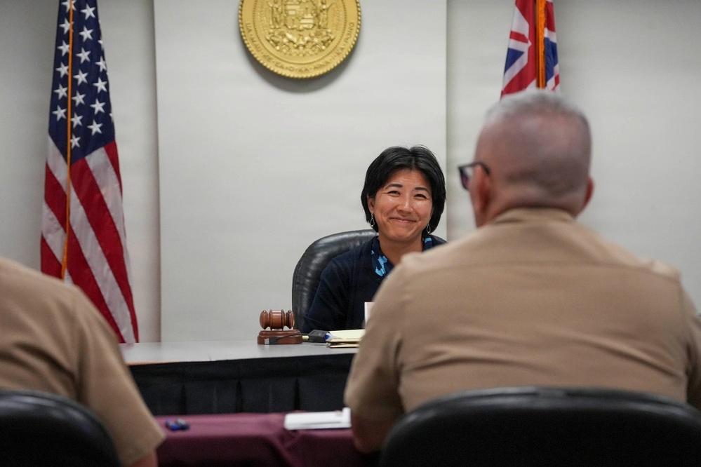 A woman seated at a judge's bench in what appears to be a courtroom smiles toward two individuals in military uniforms seated with their backs to the camera. A gavel is visible on the bench, and the setting includes an American flag and what appears to be the Hawaii state seal and flag on the wall behind her. The scene suggests a legal or official proceeding involving military personnel.