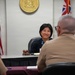 A woman seated at a judge's bench in what appears to be a courtroom smiles toward two individuals in military uniforms seated with their backs to the camera. A gavel is visible on the bench, and the setting includes an American flag and what appears to be the Hawaii state seal and flag on the wall behind her. The scene suggests a legal or official proceeding involving military personnel.