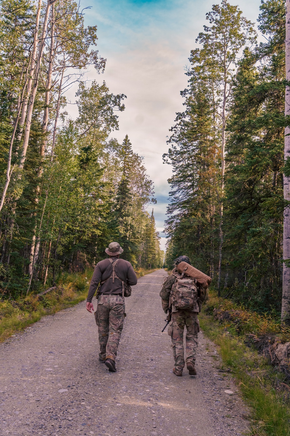 Two individuals wearing military-style camouflage uniforms and carrying tactical gear walk away from the camera down a straight gravel road flanked by tall mixed forest trees. The person on the left wears a boonie hat and carries a small hip pack, while the person on the right carries a large rucksack with a rolled sleeping pad or bedroll. The setting appears to be a remote forested area, likely in a northern or boreal region based on the tree species visible.