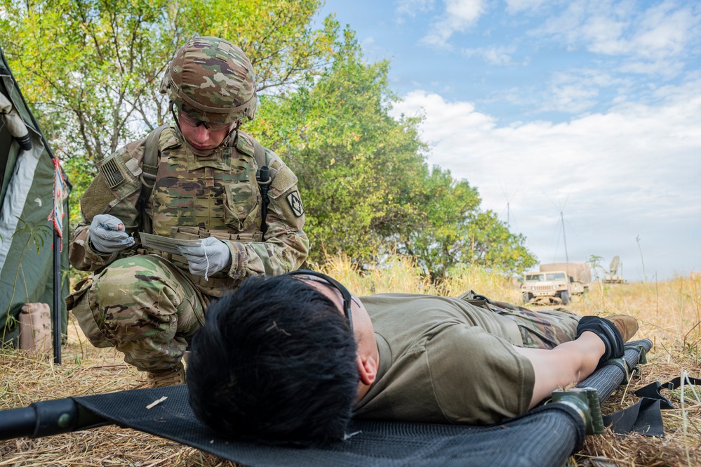 A U.S. Army soldier in full OCP (Operational Camouflage Pattern) uniform and combat gear is kneeling over a person lying face-down on a litter/stretcher in an outdoor field setting. The soldier is wearing medical gloves and appears to be reviewing a document or casualty card, suggesting a tactical combat casualty care (TCCC) training exercise. A military tent, vegetation, and a HMMWV (Humvee) are visible in the background.