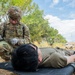 A U.S. Army soldier in full OCP (Operational Camouflage Pattern) uniform and combat gear is kneeling over a person lying face-down on a litter/stretcher in an outdoor field setting. The soldier is wearing medical gloves and appears to be reviewing a document or casualty card, suggesting a tactical combat casualty care (TCCC) training exercise. A military tent, vegetation, and a HMMWV (Humvee) are visible in the background.