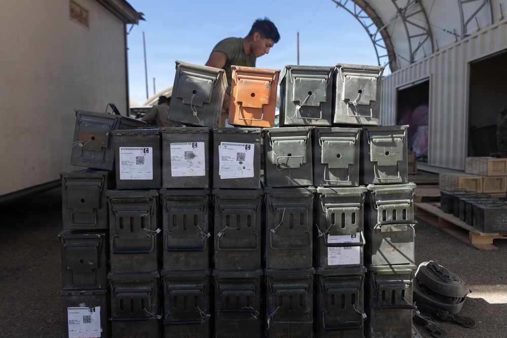 A person in a military-style t-shirt is stacking or organizing olive drab metal ammunition cans in an outdoor military logistics area. The cans are arranged in a large, organized stack and bear white labels with QR codes and text markings. The setting appears to be a forward operating base or supply depot with a Quonset-style structure visible in the background.