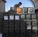 A person in a military-style t-shirt is stacking or organizing olive drab metal ammunition cans in an outdoor military logistics area. The cans are arranged in a large, organized stack and bear white labels with QR codes and text markings. The setting appears to be a forward operating base or supply depot with a Quonset-style structure visible in the background.