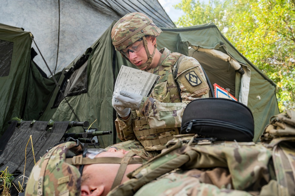 A U.S. Army soldier wearing OCP (Operational Camouflage Pattern) uniform and combat gear is reviewing what appears to be a medical casualty card or field medical form while kneeling over a fellow soldier lying on the ground. The scene takes place in a field training environment near a military tent, suggesting a combat casualty care or medical training exercise. The standing soldier is wearing latex gloves, indicating active medical treatment or assessment.