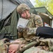 A U.S. Army soldier wearing OCP (Operational Camouflage Pattern) uniform and combat gear is reviewing what appears to be a medical casualty card or field medical form while kneeling over a fellow soldier lying on the ground. The scene takes place in a field training environment near a military tent, suggesting a combat casualty care or medical training exercise. The standing soldier is wearing latex gloves, indicating active medical treatment or assessment.