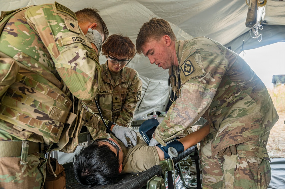 Three U.S. Army soldiers in OCP (Operational Camouflage Pattern) uniforms are providing medical treatment to a patient lying on a military litter inside a field tent. The personnel, identifiable by name tapes 'SIDES' and 'RAMIREZ', are performing what appears to be a medical assessment including blood pressure measurement and airway management. This appears to be either a field training exercise or an actual forward medical treatment scenario.
