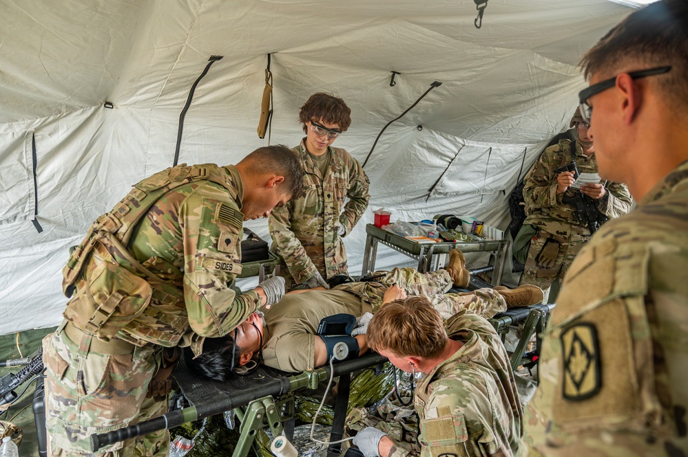 Military personnel in OCP (Operational Camouflage Pattern) uniforms are conducting a medical training exercise inside a field tent. Several soldiers are treating a simulated casualty lying on a military litter/stretcher, with one soldier checking vitals using a blood pressure cuff while another attends to the patient's upper body. Additional soldiers observe or assist in the background, suggesting a combat medic training scenario.