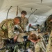 Military personnel in OCP (Operational Camouflage Pattern) uniforms are conducting a medical training exercise inside a field tent. Several soldiers are treating a simulated casualty lying on a military litter/stretcher, with one soldier checking vitals using a blood pressure cuff while another attends to the patient's upper body. Additional soldiers observe or assist in the background, suggesting a combat medic training scenario.