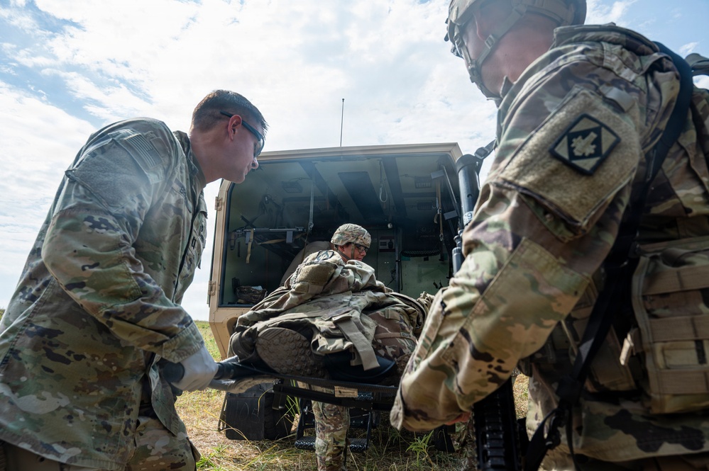 Military personnel in U.S. Army OCP (Operational Camouflage Pattern) uniforms are conducting a medical evacuation or casualty handling exercise near a military vehicle. Two soldiers are handling a litter (stretcher) with a simulated or actual casualty being loaded or unloaded from what appears to be a military ambulance or medical vehicle. The setting appears to be an open field during daylight training conditions.
