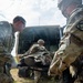 Military personnel in U.S. Army OCP (Operational Camouflage Pattern) uniforms are conducting a medical evacuation or casualty handling exercise near a military vehicle. Two soldiers are handling a litter (stretcher) with a simulated or actual casualty being loaded or unloaded from what appears to be a military ambulance or medical vehicle. The setting appears to be an open field during daylight training conditions.