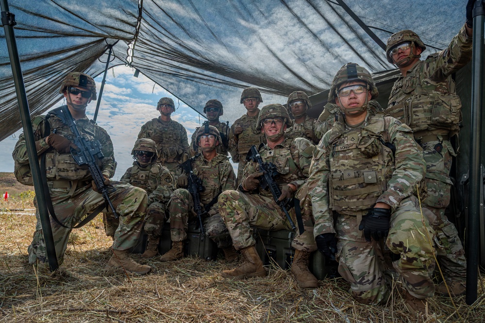 A group of approximately ten U.S. Army soldiers in full combat gear pose together under a camouflage netting canopy in an outdoor field environment. The soldiers are wearing Operational Camouflage Pattern (OCP) uniforms, helmets, plate carriers, and are armed with M4-style carbines. The setting appears to be a training exercise or field operation based on the temporary shelter and dry grassland terrain.