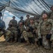 A group of approximately ten U.S. Army soldiers in full combat gear pose together under a camouflage netting canopy in an outdoor field environment. The soldiers are wearing Operational Camouflage Pattern (OCP) uniforms, helmets, plate carriers, and are armed with M4-style carbines. The setting appears to be a training exercise or field operation based on the temporary shelter and dry grassland terrain.