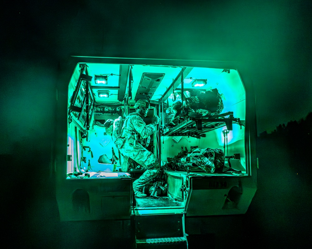 A military medical personnel in camouflage uniform is working inside what appears to be a military ambulance or medical vehicle at night. The interior is illuminated by distinctive green lighting, likely tactical/blackout-compatible medical lighting used to preserve night vision. At least one patient or additional personnel appears to be present on a stretcher or litter inside the vehicle.