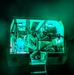 A military medical personnel in camouflage uniform is working inside what appears to be a military ambulance or medical vehicle at night. The interior is illuminated by distinctive green lighting, likely tactical/blackout-compatible medical lighting used to preserve night vision. At least one patient or additional personnel appears to be present on a stretcher or litter inside the vehicle.