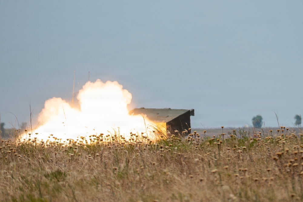 The image shows a large explosion or weapons discharge occurring in an open field, with intense white and orange flames and smoke billowing upward. A dark rectangular structure, possibly a military vehicle or bunker, is visible to the right of the blast. The foreground features dry grassland with wildflowers, suggesting a rural or training range environment.