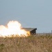 The image shows a large explosion or weapons discharge occurring in an open field, with intense white and orange flames and smoke billowing upward. A dark rectangular structure, possibly a military vehicle or bunker, is visible to the right of the blast. The foreground features dry grassland with wildflowers, suggesting a rural or training range environment.