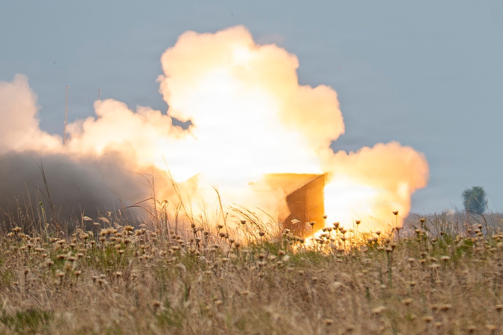 The image shows a large explosion or fire event occurring in an open field with dry grassland and wildflowers in the foreground. Intense orange and yellow flames are visible at the center, surrounded by billowing white and gray smoke clouds rising into a pale sky. A partially visible dark structure or vehicle appears to be engulfed in or adjacent to the blast.