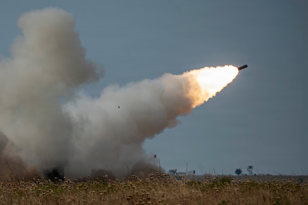 The image captures a rocket or missile being launched, with a large plume of white smoke trailing behind it as it ascends at an angle into an overcast sky. The launch appears to be occurring over an open field with dry grassland vegetation in the foreground. The projectile is clearly visible at the leading edge of the smoke trail with visible exhaust flame.