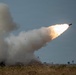 The image captures a rocket or missile being launched, with a large plume of white smoke trailing behind it as it ascends at an angle into an overcast sky. The launch appears to be occurring over an open field with dry grassland vegetation in the foreground. The projectile is clearly visible at the leading edge of the smoke trail with visible exhaust flame.