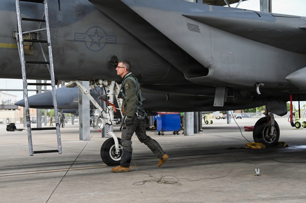 A military pilot in full flight gear conducts a pre-flight walk-around inspection of a gray fighter jet, identifiable as an F-15 Eagle based on its distinctive fuselage shape. The pilot is walking beneath the aircraft on a concrete flight line, examining the underside and landing gear components. The setting appears to be a military air base with maintenance equipment and other aircraft visible in the background.