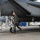 A military pilot in full flight gear conducts a pre-flight walk-around inspection of a gray fighter jet, identifiable as an F-15 Eagle based on its distinctive fuselage shape. The pilot is walking beneath the aircraft on a concrete flight line, examining the underside and landing gear components. The setting appears to be a military air base with maintenance equipment and other aircraft visible in the background.