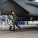 A military pilot in full flight gear conducts a pre-flight walk-around inspection of a gray fighter jet, identifiable as an F-15 Eagle based on its distinctive fuselage shape. The pilot is walking beneath the aircraft on a concrete flight line, examining the underside and landing gear components. The setting appears to be a military air base with maintenance equipment and other aircraft visible in the background.