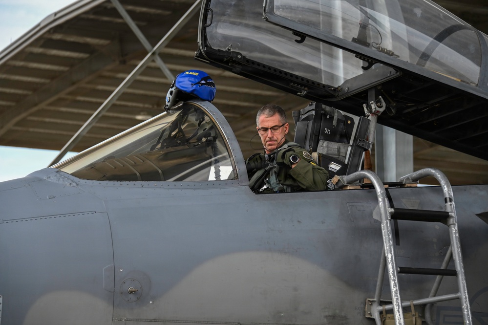 A military pilot in a green flight suit is seated in the cockpit of a gray fighter jet, appearing to conduct pre-flight checks or equipment inspection. A blue flight helmet with text visible on it rests on the front cockpit headrest. The aircraft is parked under a covered shelter structure with a maintenance ladder positioned alongside.