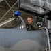 A military pilot in a green flight suit is seated in the cockpit of a gray fighter jet, appearing to conduct pre-flight checks or equipment inspection. A blue flight helmet with text visible on it rests on the front cockpit headrest. The aircraft is parked under a covered shelter structure with a maintenance ladder positioned alongside.