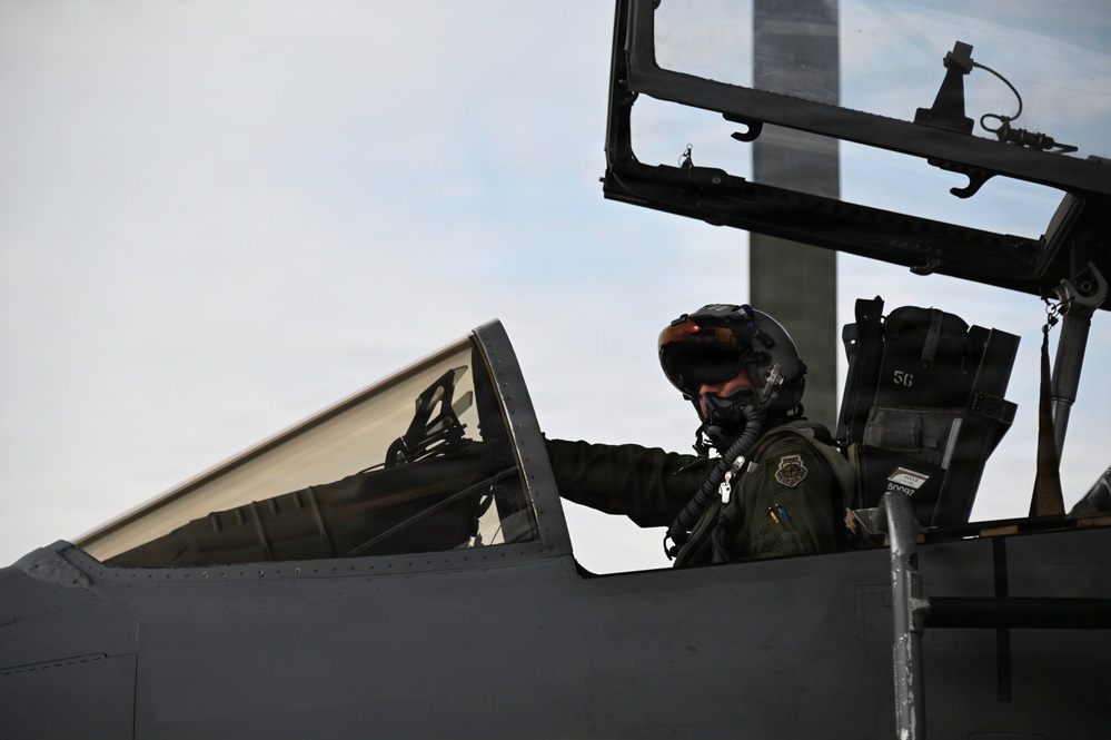 A military pilot is seated in the cockpit of a fighter jet, wearing full flight gear including a helmet with visor and oxygen mask. The canopy is open and raised above the cockpit. The aircraft appears to be a modern U.S. Air Force jet, likely an F-15 or similar platform, based on the cockpit design and gray fuselage.