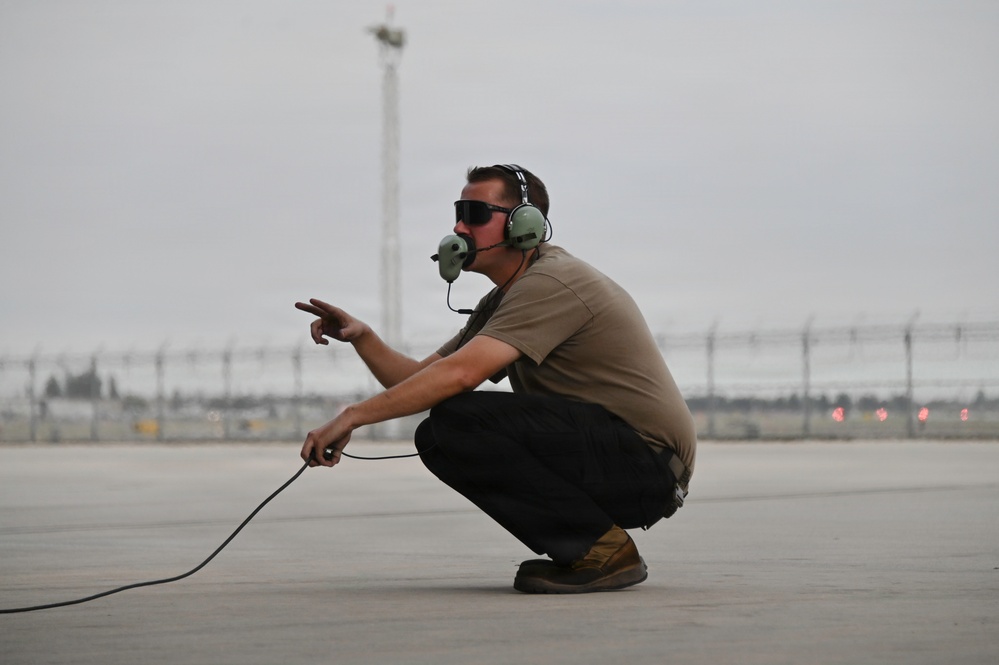 A military or aviation ground crew member is crouching on a tarmac while wearing a communications headset with a boom microphone and sunglasses. He is holding a communication cable and pointing with one hand, likely directing aircraft or communicating with a flight crew. The setting appears to be a military airfield or airport flight line.