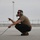 A military or aviation ground crew member is crouching on a tarmac while wearing a communications headset with a boom microphone and sunglasses. He is holding a communication cable and pointing with one hand, likely directing aircraft or communicating with a flight crew. The setting appears to be a military airfield or airport flight line.