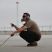A military or aviation ground crew member is crouching on a tarmac while wearing a communications headset with a boom microphone and sunglasses. He is holding a communication cable and pointing with one hand, likely directing aircraft or communicating with a flight crew. The setting appears to be a military airfield or airport flight line.