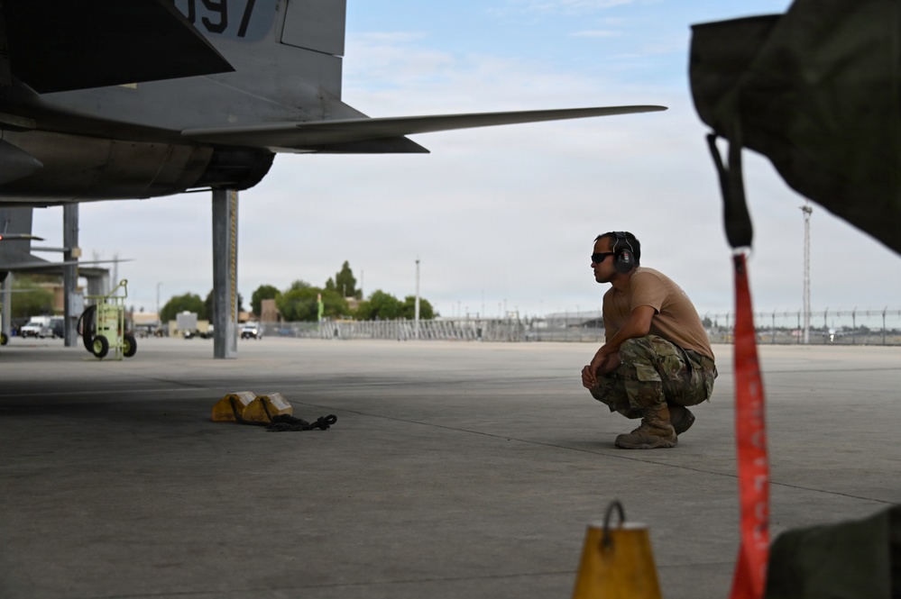 A military aircraft maintainer in OCP (Operational Camouflage Pattern) uniform crouches beneath a military jet aircraft on a flight line, appearing to conduct a pre-flight or post-flight inspection. The technician is wearing hearing protection and sunglasses while examining the underside of the aircraft. Wheel chocks and safety streamers are visible nearby, indicating standard ground safety procedures are in place.