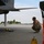 A military aircraft maintainer in OCP (Operational Camouflage Pattern) uniform crouches beneath a military jet aircraft on a flight line, appearing to conduct a pre-flight or post-flight inspection. The technician is wearing hearing protection and sunglasses while examining the underside of the aircraft. Wheel chocks and safety streamers are visible nearby, indicating standard ground safety procedures are in place.