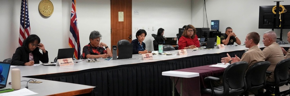 A formal meeting or briefing is taking place in what appears to be a government conference room in Hawaii, indicated by the Hawaii state flag and official seal visible on the wall. Several civilians and military personnel in tan/khaki uniforms are seated around a large conference table with name placards. The setting suggests an official government or military coordination meeting.