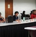 A formal meeting or briefing is taking place in what appears to be a government conference room in Hawaii, indicated by the Hawaii state flag and official seal visible on the wall. Several civilians and military personnel in tan/khaki uniforms are seated around a large conference table with name placards. The setting suggests an official government or military coordination meeting.