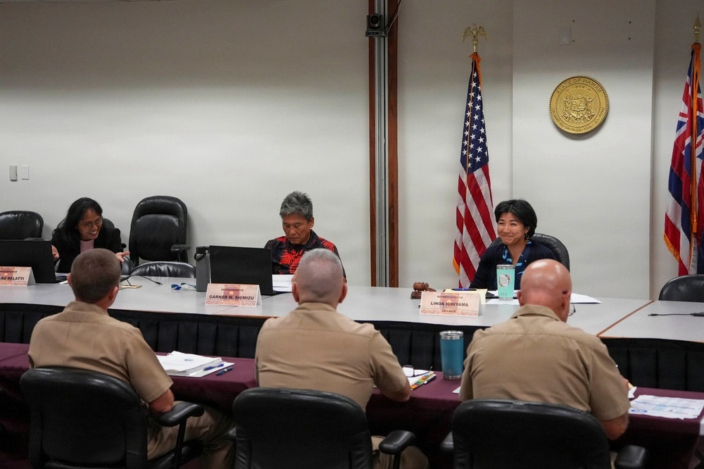 A formal meeting or hearing is taking place in what appears to be an official Hawaii government conference room, indicated by the State of Hawaii seal on the wall and both U.S. and Hawaii state flags. Civilian officials or representatives sit on one side of a conference table facing military personnel in tan/khaki uniforms seated with their backs to the camera. Name placards visible on the table identify participants including 'Garner M. Shimizu' and 'Linda Ichiyama.'