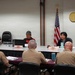 A formal meeting or hearing is taking place in what appears to be an official Hawaii government conference room, indicated by the State of Hawaii seal on the wall and both U.S. and Hawaii state flags. Civilian officials or representatives sit on one side of a conference table facing military personnel in tan/khaki uniforms seated with their backs to the camera. Name placards visible on the table identify participants including 'Garner M. Shimizu' and 'Linda Ichiyama.'