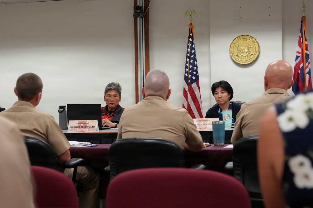 A formal government meeting is taking place in what appears to be a Hawaii State Legislature committee room. Two representatives, identified by nameplates as Garner M. Shimizu and Linda Ichiyama (Co-Chair), are seated across from several military personnel in tan uniforms. The State of Hawaii seal and both the U.S. and Hawaii state flags are prominently displayed on the wall behind the representatives.