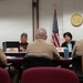 A formal government meeting is taking place in what appears to be a Hawaii State Legislature committee room. Two representatives, identified by nameplates as Garner M. Shimizu and Linda Ichiyama (Co-Chair), are seated across from several military personnel in tan uniforms. The State of Hawaii seal and both the U.S. and Hawaii state flags are prominently displayed on the wall behind the representatives.