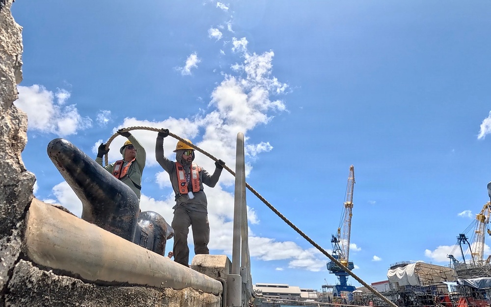Two workers in safety gear are handling a heavy rope or mooring line at what appears to be a maritime or shipyard facility. They are positioned near large mooring bollards on a dock or vessel, with industrial cranes visible in the background. The scene suggests active docking, mooring, or rigging operations at a port or naval facility.